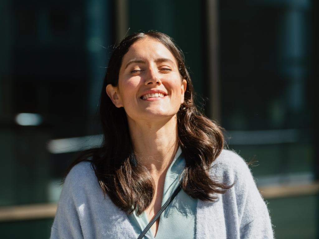 En kvinne smiler bredt utendørs og nyter solens varme mot ansiktet